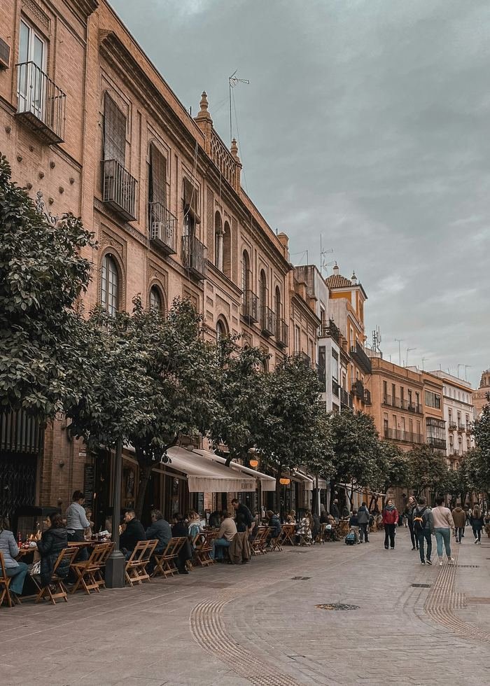 A Seville street in soft light: orange trees along a wide pavement, terrace tables outside a bar, people walking and sitting.