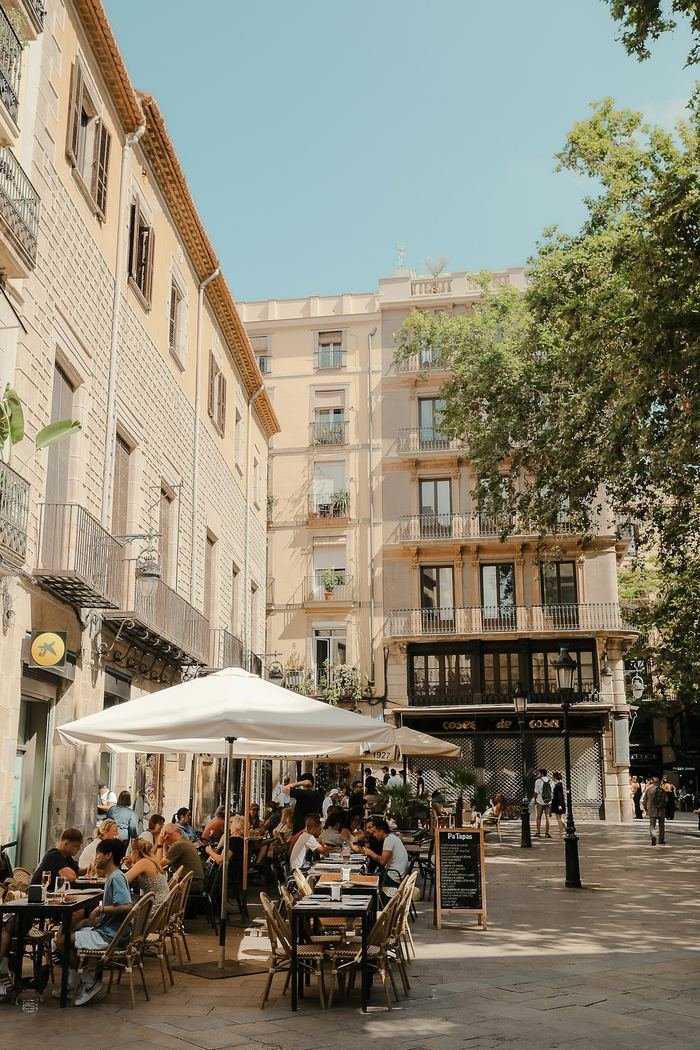A sunlit Spanish street corner: pavement café tables under a white parasol, apartment balconies above, a plane tree, people at the tables and walking past.