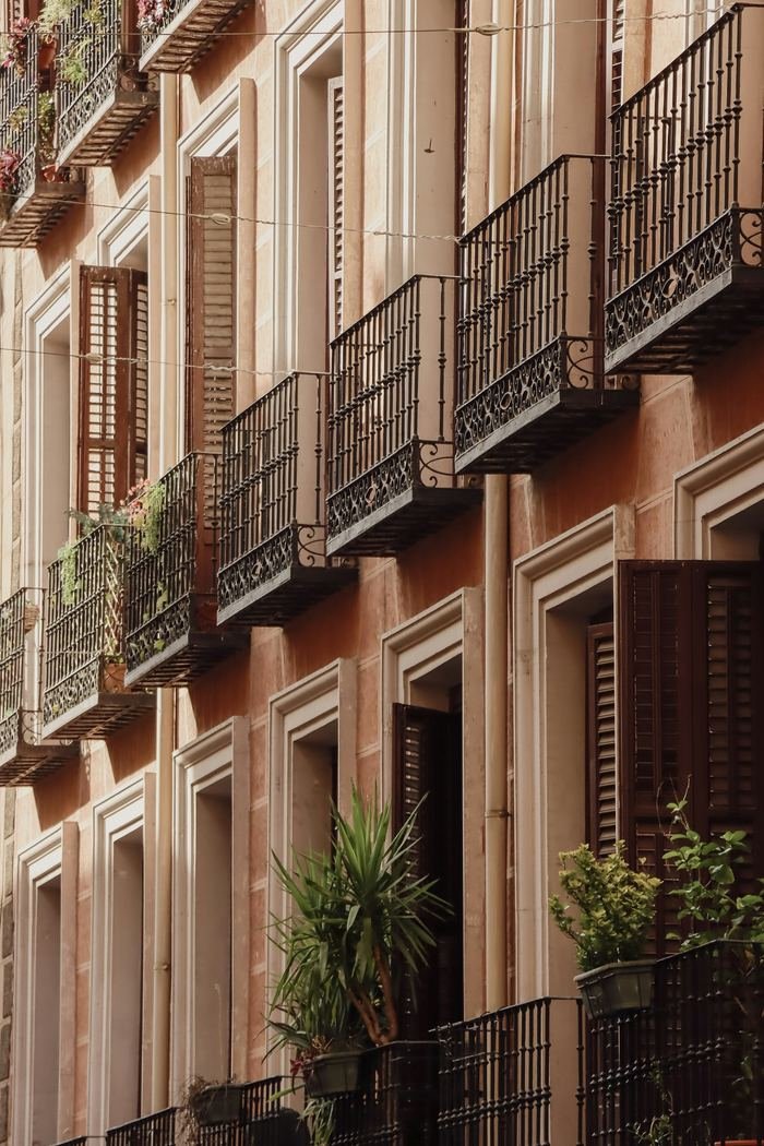 A Madrid façade in warm afternoon light — wrought-iron balconies, wooden shutters, plants in pots, one shutter open.